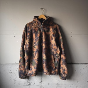 May include: A full-zip fleece jacket with a leaf pattern in shades of brown, orange, and green. The jacket has a high collar, two side pockets, and is hanging on a wooden hanger against a gray wall.
