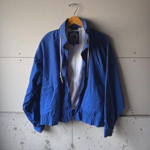 Op de afbeelding: Een helderblauwe jas met witte voering hangt aan een houten hanger. De jas heeft een volledige ritssluiting, een trekkoord bij de kraag en elastische manchetten. Er is een logopatch aan de binnenkant.