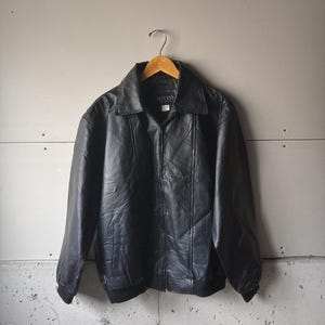 Puede incluir: Chaqueta de cuero negra con cuello y cierre de cremallera. La chaqueta tiene dobladillo y puños de canalé. La marca "BOTANY" es visible en la etiqueta. La chaqueta está colgada en una percha de madera.
