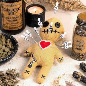 May include: A tan burlap voodoo doll featuring black button eyes, a stitched mouth, and a red felt heart on the chest. The doll is pierced by multiple silver pins topped with small white skull beads, and has black stitched X marks on limbs.
