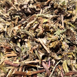May include: A collection of dried herbal plant matter consisting of fragmented green leaves, brown stems, and small woody pieces. The mixture displays a variety of earthy tones including dark forest green, olive, tan, and deep brown, with a textured appearance.