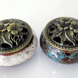 May include: Two round ceramic incense burners with removable brass-toned metal lids. The lids feature an intricate floral cutout design. One burner has a speckled white and brown glaze, while the other has a mottled blue and brown glaze finish.