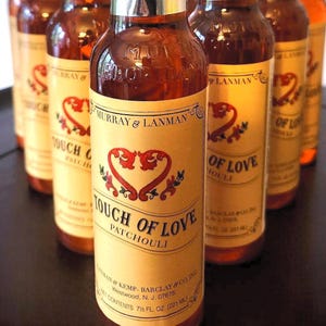 Puede incluir: Varias botellas de perfume "Touch of Love Patchouli" alineadas. Cada botella tiene una etiqueta crema con diseños de corazón rojo y el texto "MURRAY & LANMAN". Contiene 221 ml de líquido.