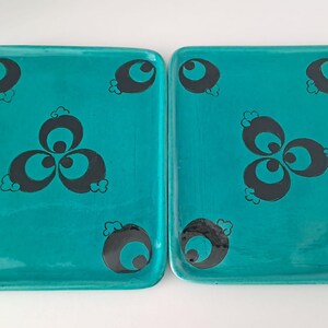 May include: Two rectangular ceramic trays with a glossy turquoise finish. Each tray features a repeating black graphic pattern consisting of a central three-lobed motif surrounded by four smaller circular elements placed near the corners.