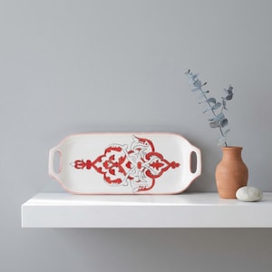 Puede incluir: Una bandeja rectangular de cerámica blanca con un diseño ornamental rojo y gris. La bandeja tiene un borde rosa y dos asas. Un pequeño jarrón de terracota con eucalipto y una pequeña piedra están junto a la bandeja.