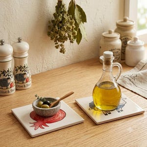 May include: Two square ceramic tiles featuring hand-painted illustrations of a red pomegranate and a black sheep. A glass cruet filled with golden olive oil sits on one tile, while a small stone bowl with olives and a wooden spoon sits on the other.