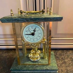 May include: A mantel clock featuring a green marble base and top, connected by four glass panels and gold-tone metal pillars. The clock face is white with black numerals and hands, positioned above a rotating gold-tone pendulum with four spheres.