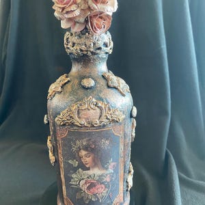 May include: Ornate, hand-painted bottle with a vintage aesthetic. The bottle features a portrait of a woman, framed by decorative elements and topped with a cluster of rose-shaped embellishments. The color palette includes bronze, black, and rose tones.