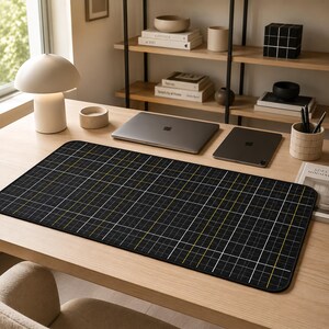 Puede incluir: Una alfombrilla de escritorio rectangular con fondo negro y un patrón de cuadrícula en blanco y amarillo. La alfombrilla tiene esquinas redondeadas y un borde cosido en negro, ofreciendo una superficie plana para portátil y tableta.