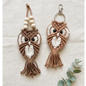 May include: Two macrame owl ornaments crafted from tan and white cord. The left owl features a hanging loop with four light wooden beads. The right owl includes a silver metal keyring attachment. Both owls have black bead eyes and fringed tails.