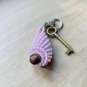 May include: A light pink macrame keychain featuring a teardrop-shaped knotted design with a single dark brown wooden bead at the base. The macrame piece is attached to a silver-toned metal split ring and a vintage-style bronze-toned skeleton key.
