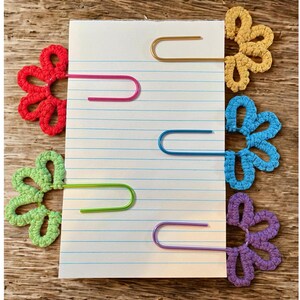 May include: Five metal paper clips, each featuring a colorful crocheted flower topper. The set includes one red, one light green, one gold, one blue, and one purple flower attached to standard-sized wire paper clips, clipped onto a lined notepad.