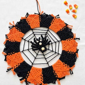 May include: A circular macrame wall hanging featuring alternating black and orange knotted sections forming a border around a central black spider web. A black macrame spider with two light-colored wooden beads sits in the center of the web.