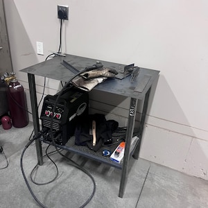 May include: A metal welding table with a black welding machine, gloves, and tools. The table has a metal frame and a shelf. A gas tank is visible in the background.