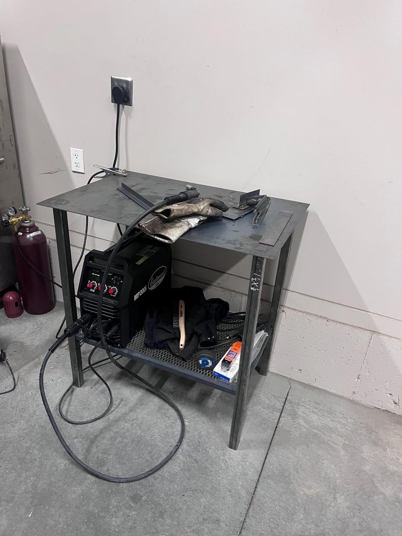 May include: A metal welding table with a black welding machine, gloves, and tools. The table has a metal frame and a shelf. A gas tank is visible in the background.