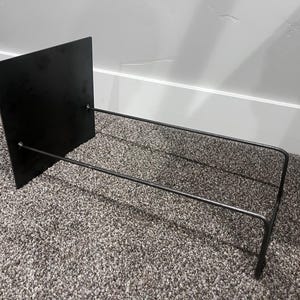 May include: A black metal book stand with a rectangular back and two horizontal bars extending forward. The stand is designed to hold books or other items. The stand is made of metal and has a minimalist design.