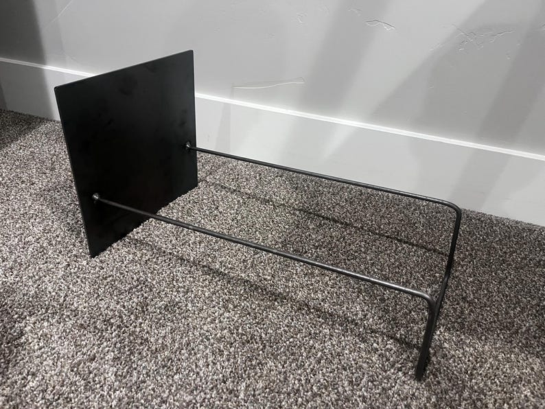 May include: A black metal book stand with a rectangular back and two horizontal bars extending forward. The stand is designed to hold books or other items. The stand is made of metal and has a minimalist design.