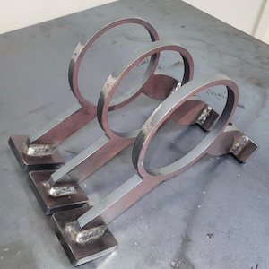 May include: A metal wine rack with three individual holders. Each holder has a circular top and a rectangular base, connected by a flat bar. The metal has a raw, industrial look with visible weld marks.