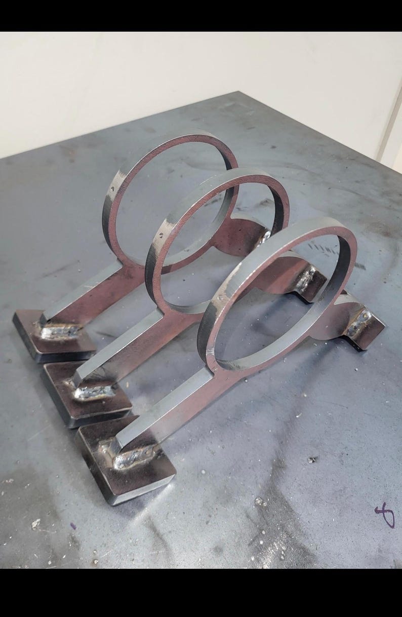 May include: A metal wine rack with three individual holders. Each holder has a circular top and a rectangular base, connected by a flat bar. The metal has a raw, industrial look with visible weld marks.