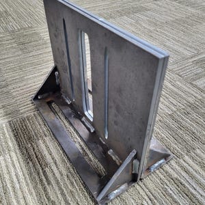 May include: A welded metal bracket with a rectangular plate featuring three vertical slots. The bracket is supported by a base and angled supports, all made of the same unfinished steel. The piece is designed for structural support.
