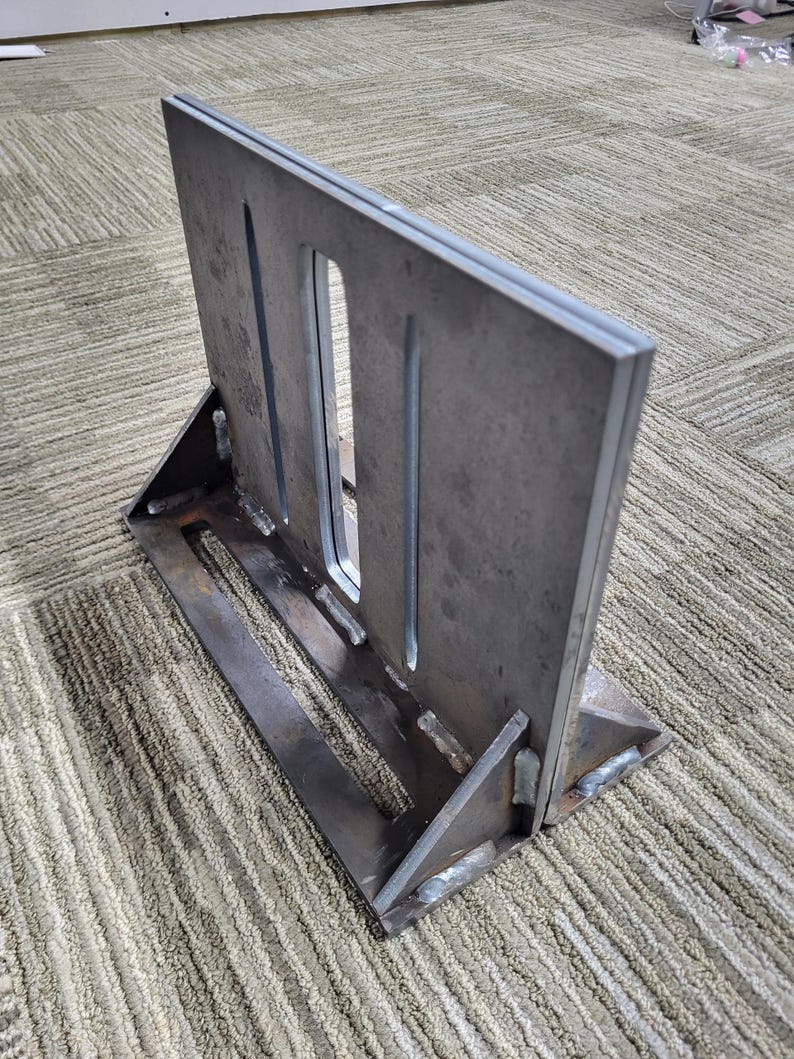 May include: A welded metal bracket with a rectangular plate featuring three vertical slots. The bracket is supported by a base and angled supports, all made of the same unfinished steel. The piece is designed for structural support.