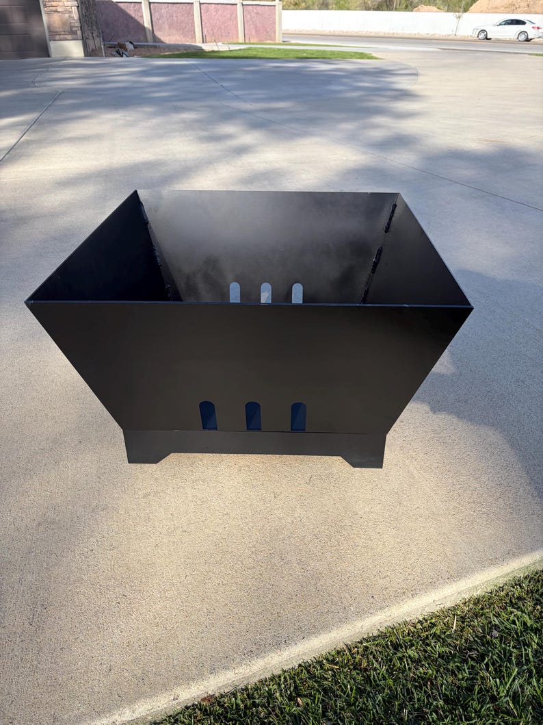 May include: A black, geometric fire pit with a square shape and angled sides. The fire pit has three rectangular cutouts on each side and sits on four small feet. Smoke is visible inside the pit.