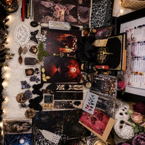 May include: A collection of dark-themed stationery items including various printed cards, stickers, bookmarks, and a journal with a gold compass design. The set features floral, celestial, and gothic motifs, alongside pens, a black scrunchie, and decorative paper elements arranged on a textured surface.