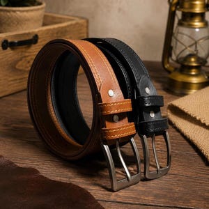 May include: Two coiled belts, one in tan and one in black, each featuring a silver-toned metal buckle, two matching belt loops, and visible stitching along the edges. The belts are positioned upright, showcasing their circular shape and hardware.