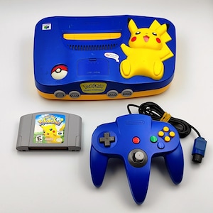 Puede incluir: Una consola de juegos Nintendo 64 azul y amarilla con un diseño de Pikachu, un mando a juego y un cartucho de juego "Pokémon Pikachu". La consola presenta una figura de Pikachu amarilla y el logotipo de "Pokémon".