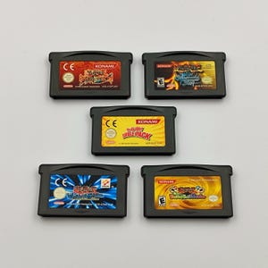 Puede incluir: Cinco cartuchos negros de Game Boy Advance. Los cartuchos tienen etiquetas coloridas con texto japonés y el logotipo de Konami. Una etiqueta dice "Yu-Gi-Oh! Double Pack". Los cartuchos están dispuestos en un patrón circular.