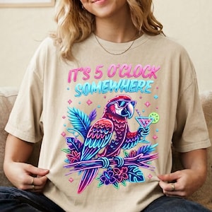 Puede incluir: Una camiseta beige con el gráfico de un loro con gafas de sol, posado en una rama con hojas y flores tropicales. El loro sostiene una copa de cóctel con una rodaja de lima. El texto dice IT'S 5 O'CLOCK SOMEWHERE en colores de neón.