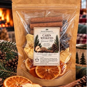 May include: A brown stand-up pouch containing a simmer pot kit labeled Cabin Weekend by Sacred Ground Co. The clear window reveals dried orange slices, apple slices, pinecones, and cinnamon sticks. The label lists scent notes of woods, orange, apple, cinnamon, and herbs, with instructions to add water and simmer on a stove.