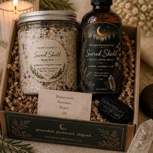 May include: A Protection Ritual Bundle in a box, featuring a jar of bath salts, a room spray bottle, a black stone, and a card with text. The products are labeled "Sacred Shield" and include the text "Protection, Presence, Peace."