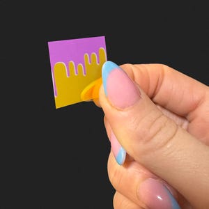 Op de afbeelding: Een kleine, vierkante sticker met een paarse bovenkant en een gele onderkant, met een druipend ontwerp. De sticker wordt vastgehouden door een hand met roze nagels en blauwe punten tegen een zwarte achtergrond.