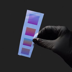 Puede incluir: Una tira de papel con cuatro cuadrados de gradiente de color, sostenida por una mano enguantada. Los cuadrados hacen la transición del púrpura al rojo y al azul. El fondo es negro sólido.