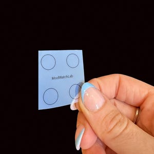 May include: A small, square, light blue translucent sticker sheet featuring four black circular outlines arranged in a grid pattern. The text ModWatchLab is printed in black in the center of the sticker sheet between the circular outlines.