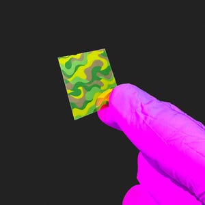 Op de afbeelding: Een klein, rechthoekig vel met een camouflagepatroon in groen, geel en bruin wordt tegen een zwarte achtergrond gehouden. Het vel wordt vastgehouden door een gehandschoende hand, met de handschoen in een felroze kleur.