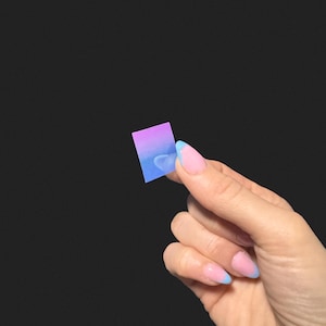 Op de afbeelding: Een klein, rechthoekig object met een verloop van paars naar blauw wordt vastgehouden door een hand met roze en blauwe French-tip nagels. De achtergrond is effen zwart.