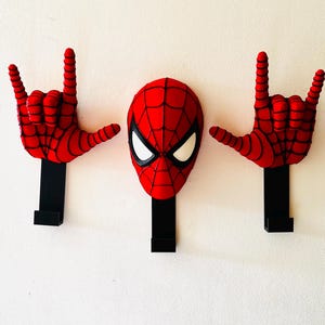 Può includere: Un set di tre ganci da parete con una maschera di Spider-Man rossa e nera al centro e due mani rosse e nere ai lati, ognuna che fa un gesto rock. I ganci sono fissati su basi rettangolari nere.