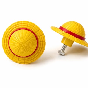 Op de afbeelding: Twee gele kastknoppen in de vorm van een strohoed, met een rode band. Eén knop ligt plat, de andere staat rechtop en toont de bout en moer voor de installatie. De knoppen staan op een witte achtergrond.