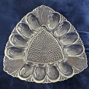 May include: Clear glass deviled egg serving dish with a triangular shape and scalloped edges. The dish has individual egg compartments and a central diamond-patterned area. The design is ornate and vintage, suitable for serving appetizers.