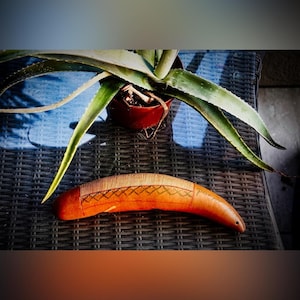 Puede incluir: Un instrumento de percusión guiro de madera curvada en color naranja-marrón, con una serie de crestas paralelas en la superficie superior y un patrón decorativo de cruz tallado a mano horizontalmente en el centro del cuerpo.