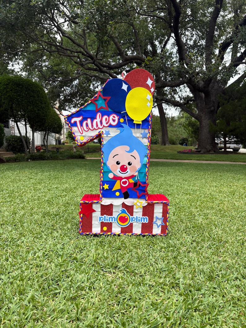 Puede incluir: Una gran decoraci&oacute;n con forma de n&uacute;mero uno que presenta el nombre Tadeo en letra blanca. El dise&ntilde;o incluye un personaje de dibujos animados azul, rayas verticales rojas y blancas y globos coloridos. La base es roja con el texto plim plim.