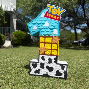 May include: A number one shaped pi&ntilde;ata featuring a Toy Story theme. The top section is light blue with white cloud cutouts, the middle is yellow with a red grid and sheriff badges, and the base is white with black cow print spots.