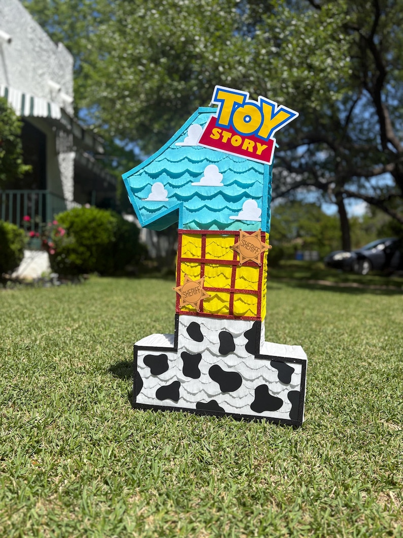 May include: A number one shaped pi&ntilde;ata featuring a Toy Story theme. The top section is light blue with white cloud cutouts, the middle is yellow with a red grid and sheriff badges, and the base is white with black cow print spots.