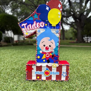 Puede incluir: Una pi&ntilde;ata con forma de n&uacute;mero uno que presenta un personaje de dibujos animados azul con nariz roja, el nombre Tadeo y el texto Plim Plim. El dise&ntilde;o incluye rayas rojas, blancas y azules, globos brillantes y peque&ntilde;os recortes de estrellas.