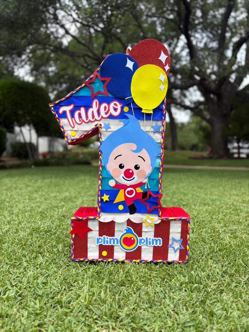 Puede incluir: Una pi&ntilde;ata con forma de n&uacute;mero uno que presenta un personaje de dibujos animados azul con nariz roja, el nombre Tadeo y el texto Plim Plim. El dise&ntilde;o incluye rayas rojas, blancas y azules, globos brillantes y peque&ntilde;os recortes de estrellas.