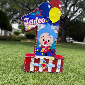 Puede incluir: Una pi&ntilde;ata con forma de n&uacute;mero uno con base de rayas rojas y blancas y detalles brillantes. Muestra el nombre Tadeo en letra blanca, un personaje de dibujos animados con pelo azul y el texto Plim Plim. Tres globos en azul, amarillo y rojo arriba.