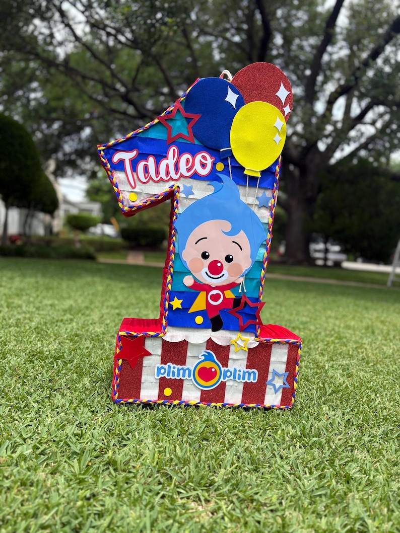 Puede incluir: Una pi&ntilde;ata con forma de n&uacute;mero uno con base de rayas rojas y blancas y detalles brillantes. Muestra el nombre Tadeo en letra blanca, un personaje de dibujos animados con pelo azul y el texto Plim Plim. Tres globos en azul, amarillo y rojo arriba.