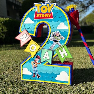 May include: A number two shaped pi&ntilde;ata featuring a blue sky background with white clouds, the Toy Story logo, and character cutouts of Buzz Lightyear and Woody. A banner hanging across the front displays the name NOAH in individual flag letters.